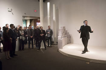 A man holding a laptop speaks in front a crowd at the launch event 'Rebooting the Greek Language'